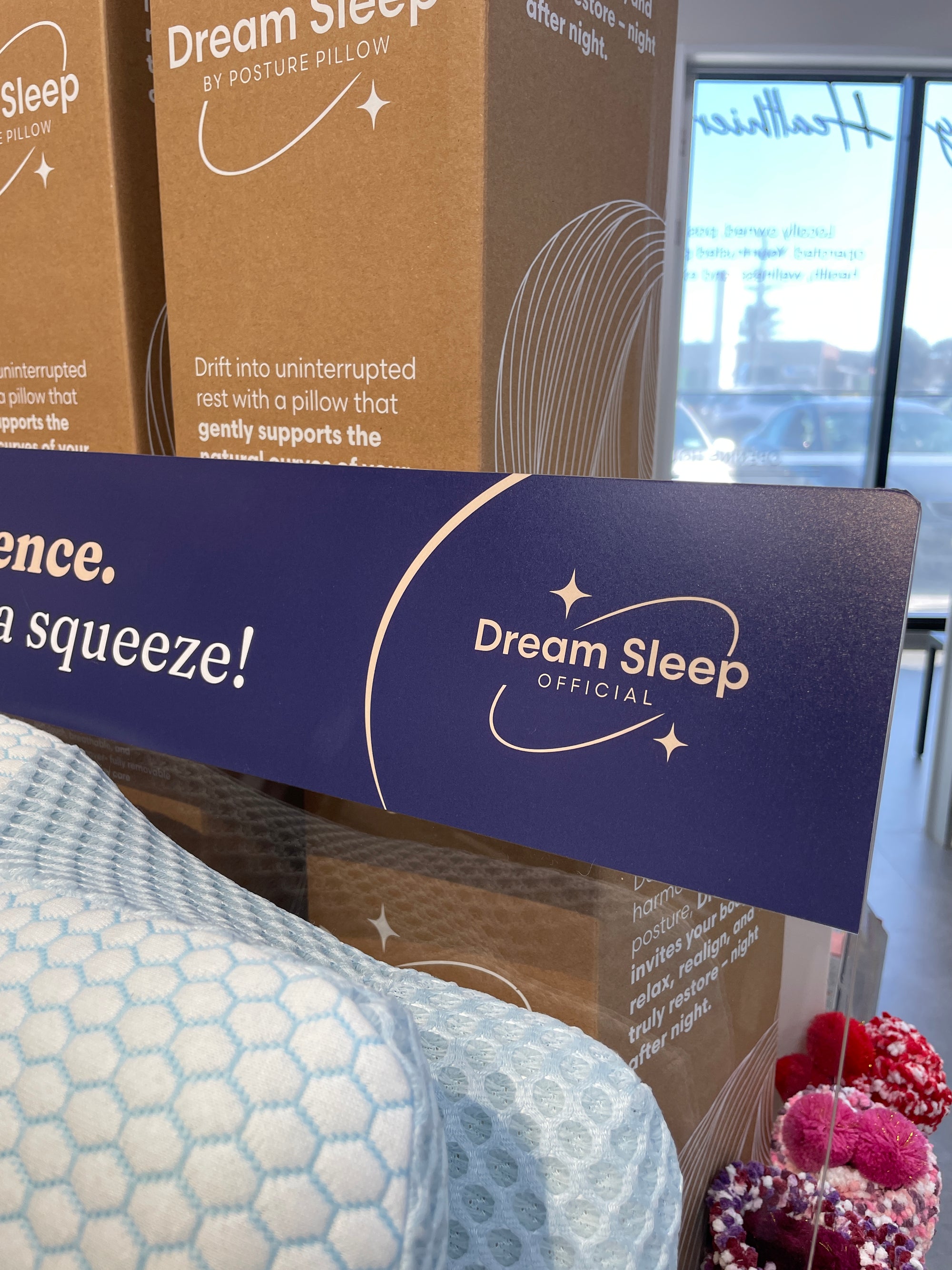 A close-up of a blue and white Dream Sleep Official Orthopaedic pillow in the foreground, with brown cardboard boxes labeled Dream Sleep behind it and a blue branded sign across the frame. A real indoor retail environment is visible through the window in the background.