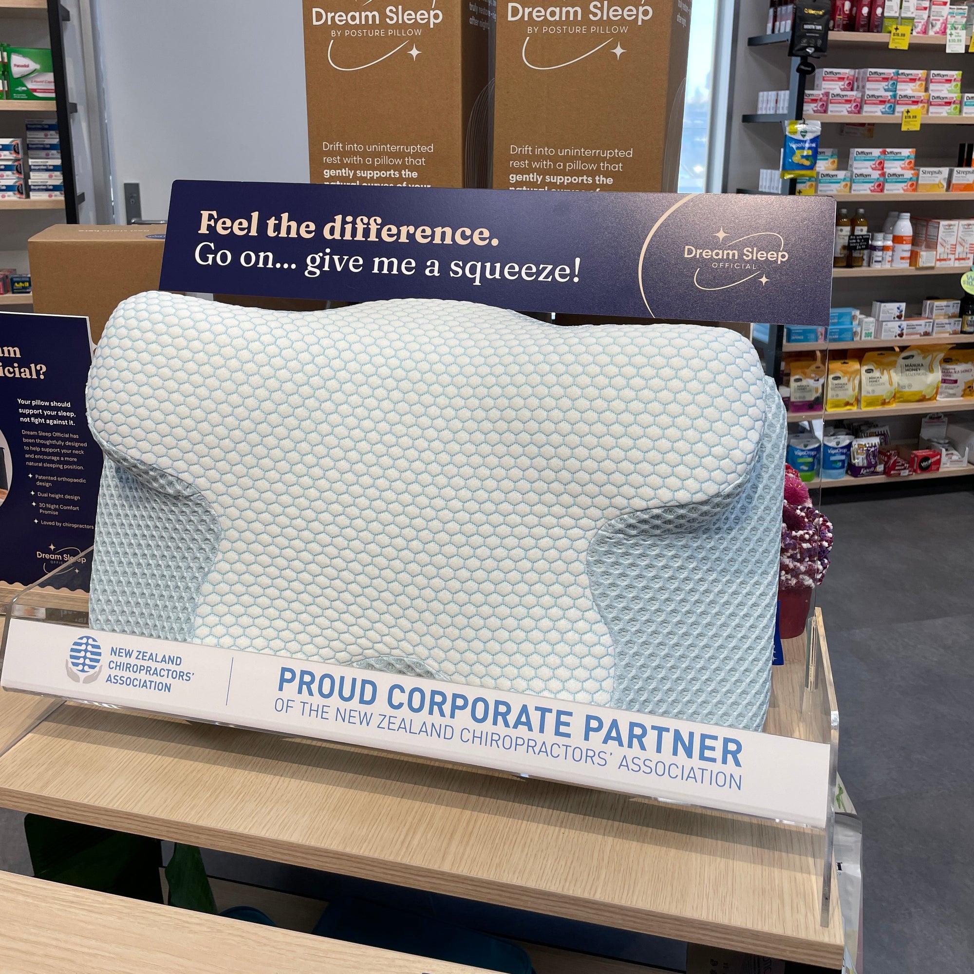 A light blue textured memory foam pillow displayed on a wooden counter inside a retail store, with Dream Sleep Official Posture Pillow NZ branded cardboard boxes and promotional signs behind it.