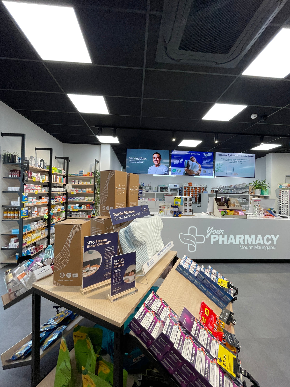 A retail pharmacy interior with shelves showcasing the Dream Sleep Pillow on the , display tables, and a counter labeled 'Your Pharmacy Mount Morgan' under ceiling lights.