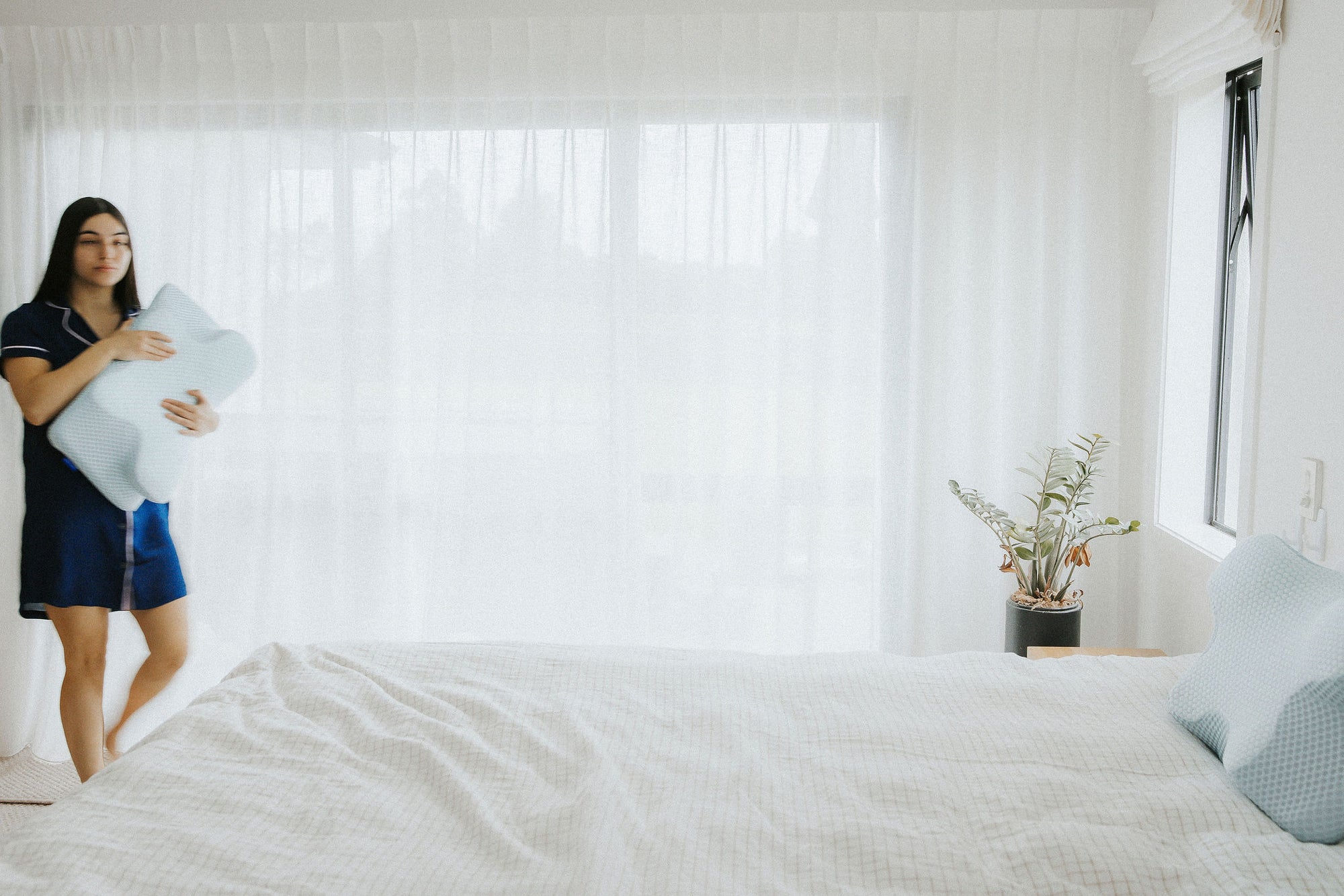 A woman holding an orthopaedic posture pillow in a bright, modern bedroom with light decor.